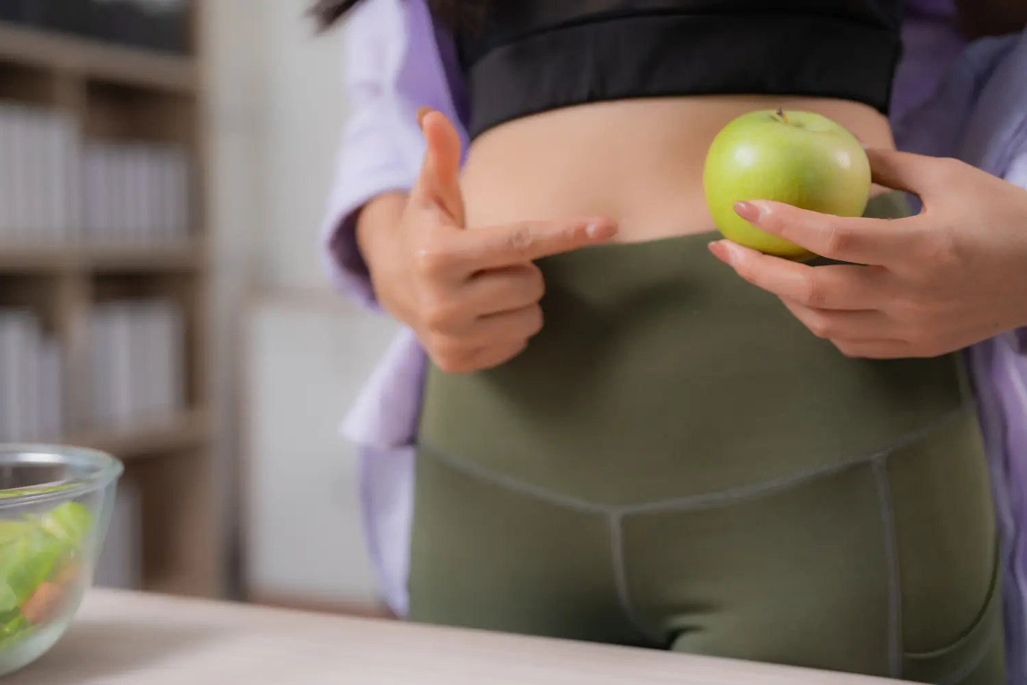 Frau in Sportkleidung zeigt auf ihren flachen Bauch und hält einen grünen Apfel in der Hand